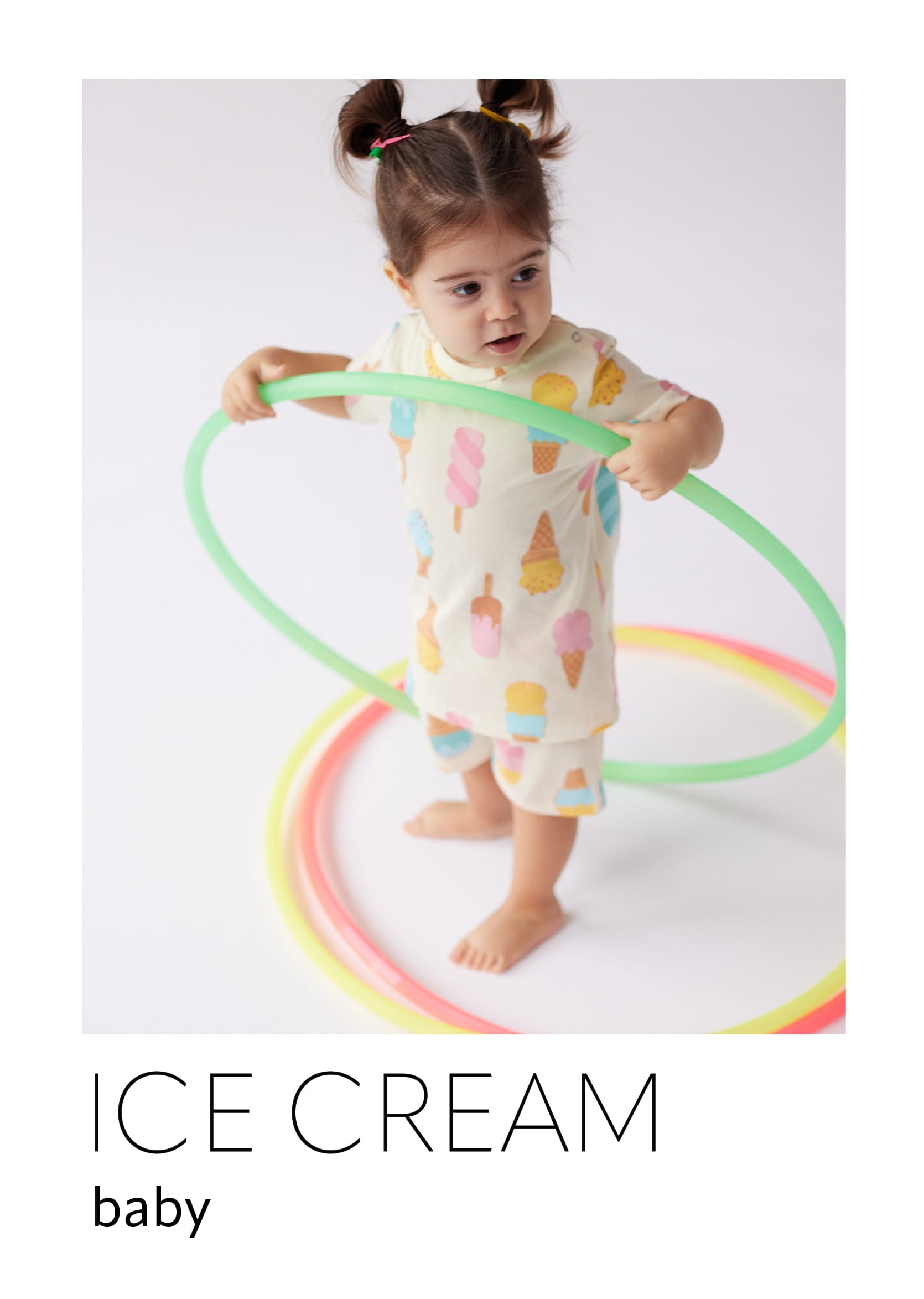 Devojčica beba stoji u beloj pidžami sa motivima sladoleda i deži zeleni obruč