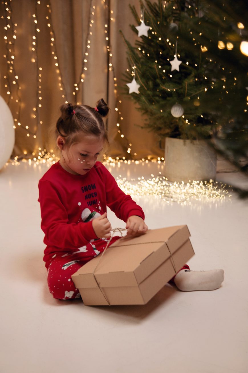 Devojcica sedi u crvenoj pidzami sedi na podu i otvara poklon