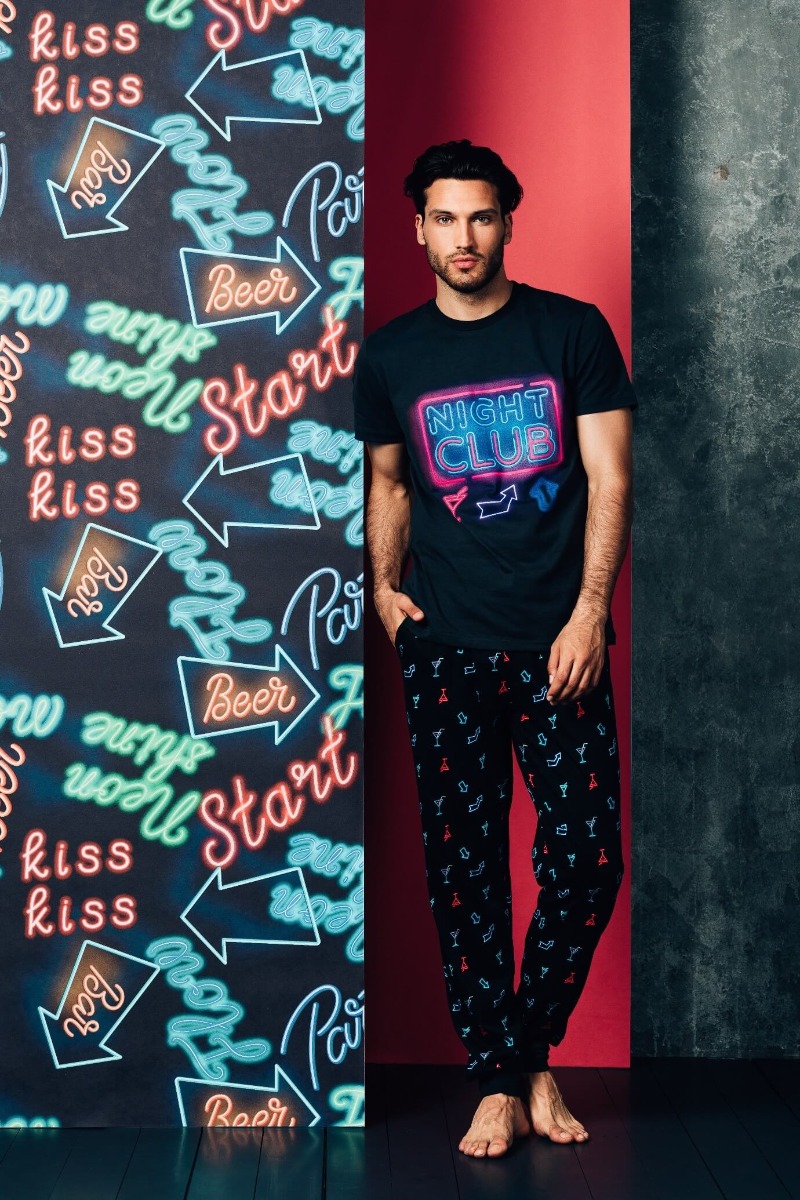 Muškarac stoji naslonjen na zid u pidžami na kratke rukave sa natpisom Night club i printom sa čašama