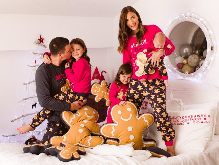 Žena i muškarac sa dve devojčice i bebom stoje na krevetu u novogodišnjim crvenim pidžamama sa motivom kolačića